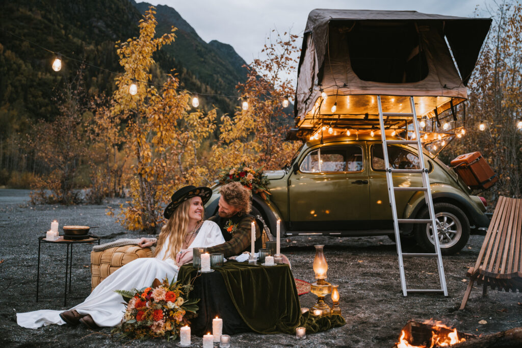 A couple sits on blankets, surrounded by candles, near a vintage car with a rooftop tent in a forested area with string lights.