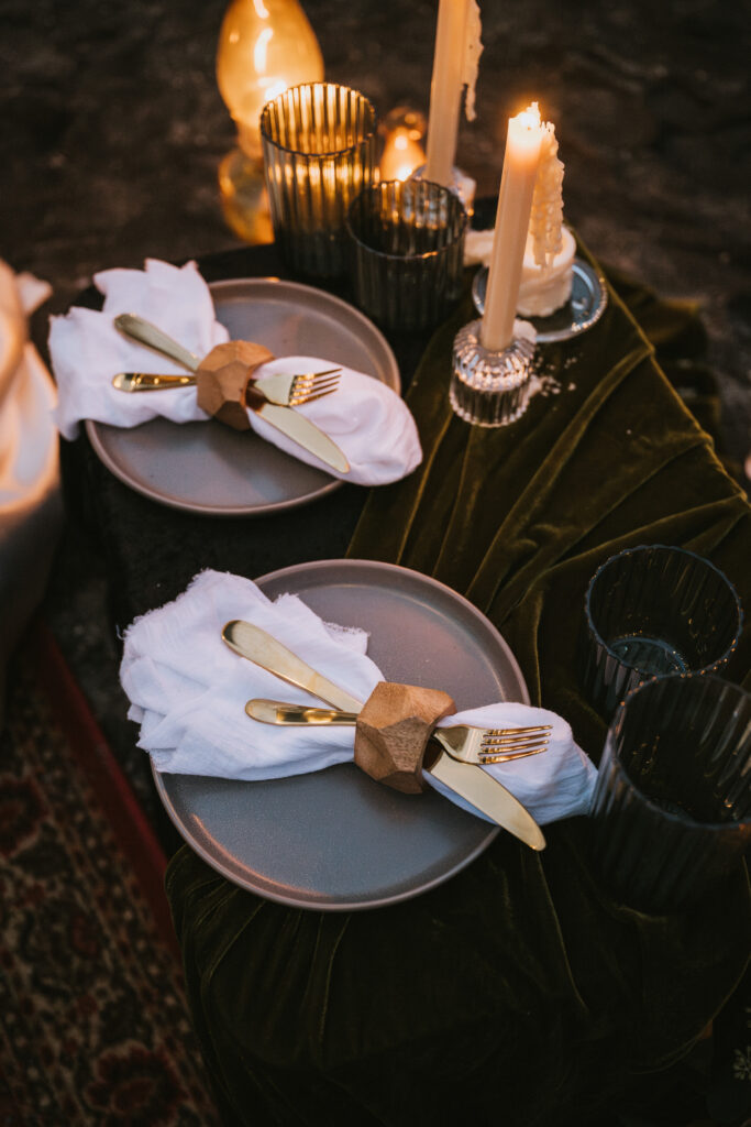 A table set for two with gray plates, golden cutlery, and white napkins. Candles and dark glass cups are arranged on a green velvet tablecloth.