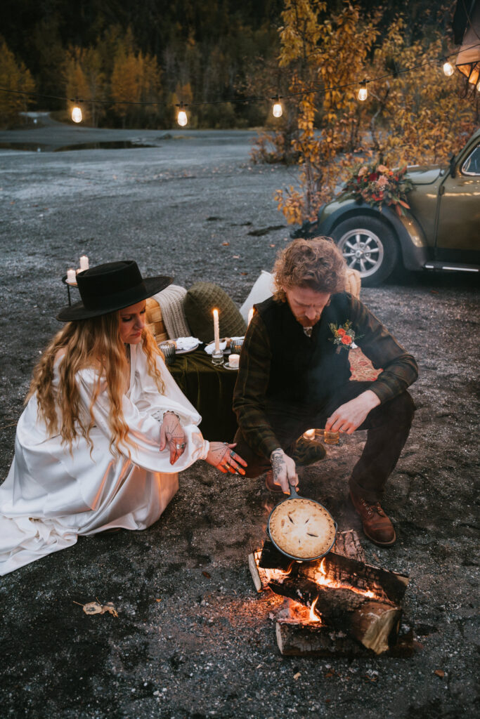 Two people sit by a campfire in a rustic outdoor setting with a pie cooking over the flames. A vintage car and a table with candles are in the background.