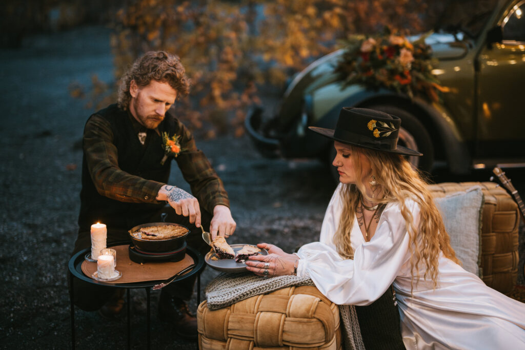 A man and woman are seated outdoors next to a small table with pies and a candle. The man is cutting a pie while the woman watches. A vintage car and autumn leaves are in the background.