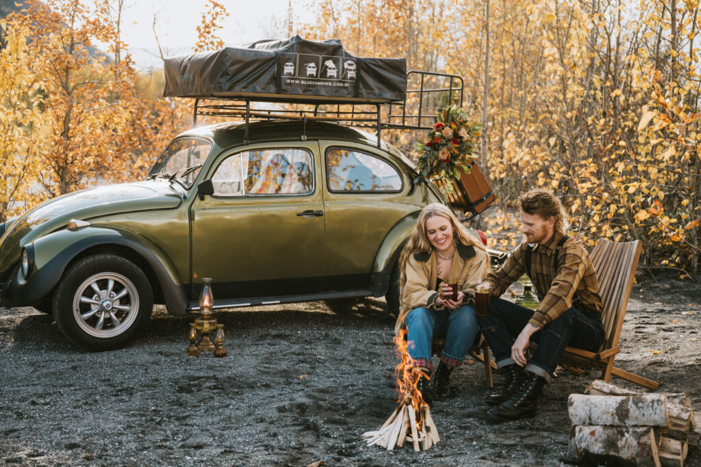 A couple sits by a campfire with a green vintage car and camping gear nearby. Autumn trees are in the background.