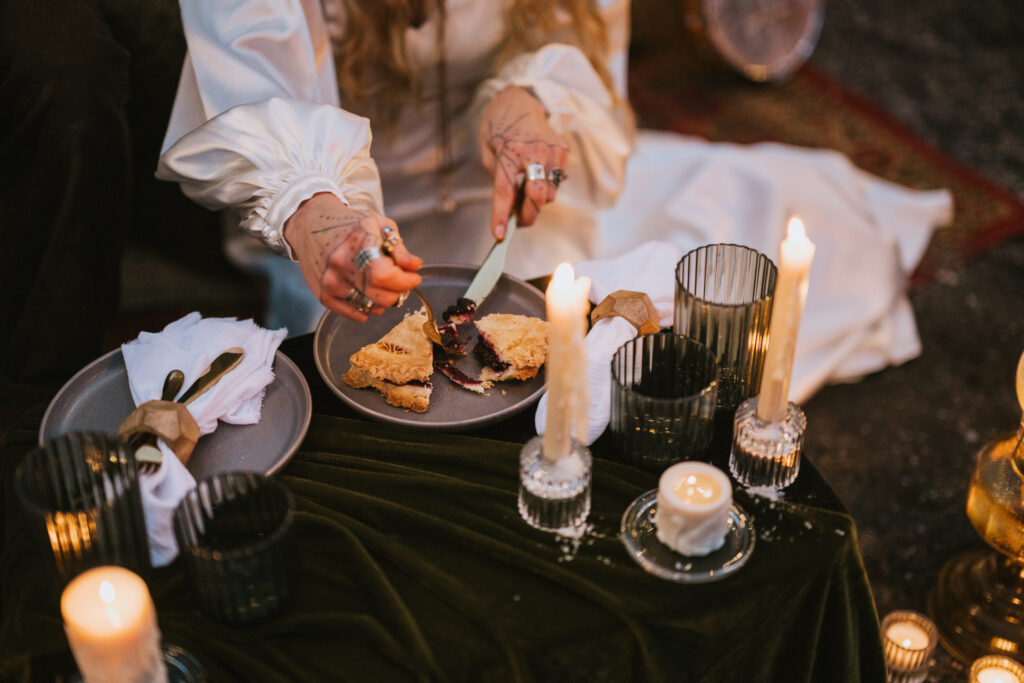 Person sitting at a candlelit table, slicing a pastry on a plate. The table is covered with a dark cloth and decorated with candles and glassware.