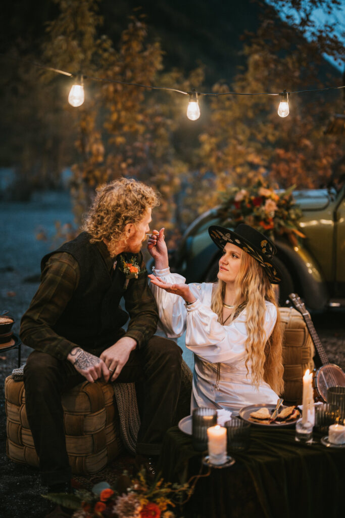 A woman in a hat and a man sit outdoors at night, under string lights. She feeds him a marshmallow. A cozy camper setup with cushions and candles is visible.