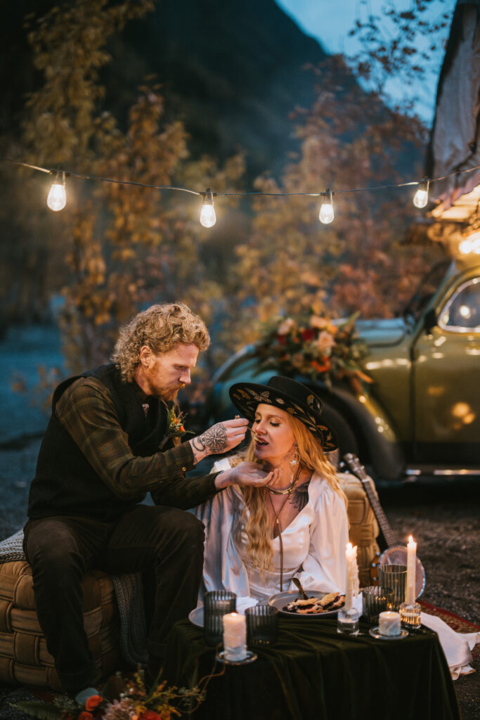 A man and woman sit together outdoors, surrounded by candles and lights. The man is feeding the woman as they sit on cushions near a classic car.