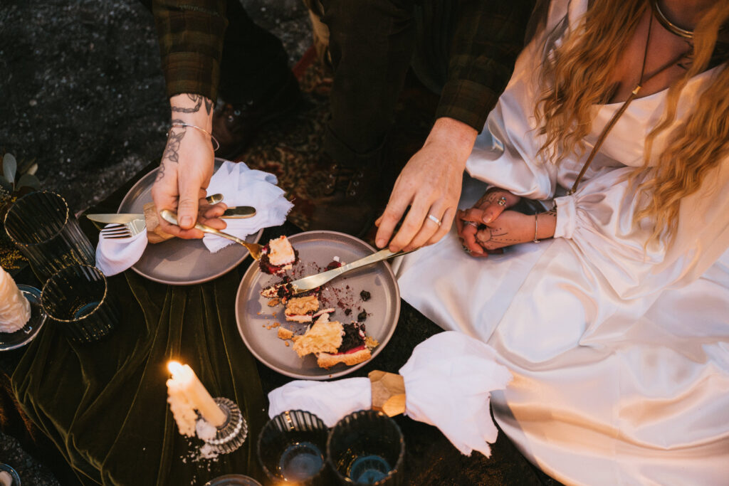 Two people share food from a plate during a picnic. One person cuts food, while the other holds a drink. Candles and dishes are on a dark cloth for a fall camping elopement