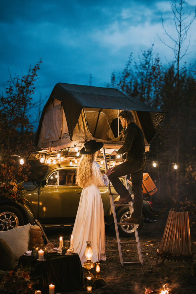 A couple sets up a rooftop tent on a vintage car surrounded by string lights and candles in a wooded area.