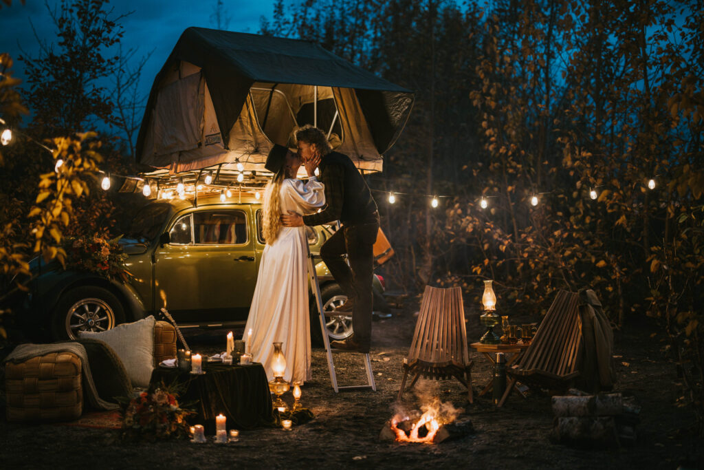 A couple embraces near a car with a rooftop tent, surrounded by string lights, chairs, a lantern, and a small campfire in a wooded area.