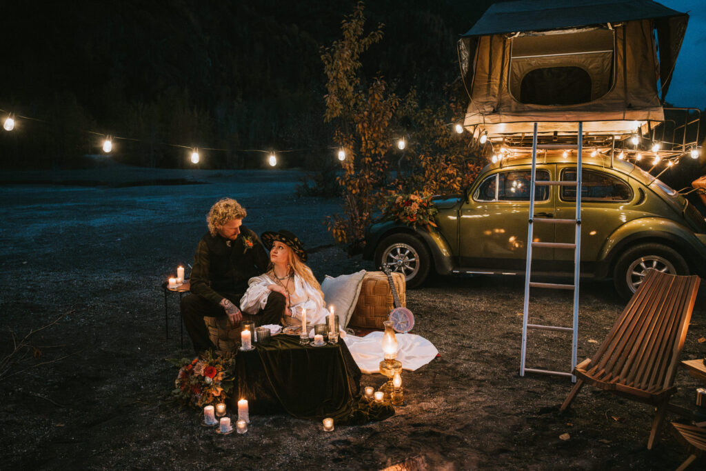 Couple sitting by candlelight with a rooftop tent on a green car in the background, surrounded by decorative items and string lights. During their relaxed elopement timeline.