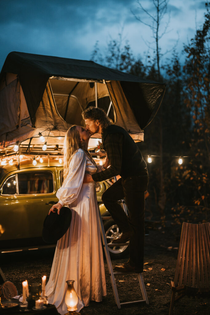 A couple kisses beside a vintage car with a rooftop tent at dusk. String lights hang around, and candles illuminate the scene.