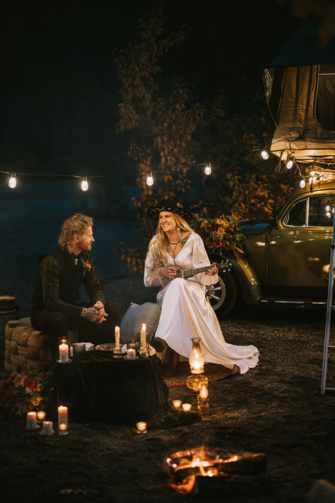 A couple sits by a campfire at night. The woman plays a ukulele, surrounded by string lights and candles. A classic car with a roof tent is in the background.