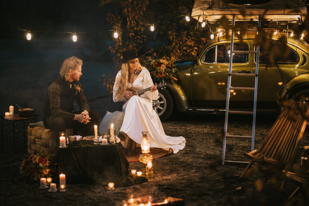 A woman plays guitar near a lantern-lit campsite beside a vintage car. A man sits across from her, surrounded by candles, with a string of lights overhead.