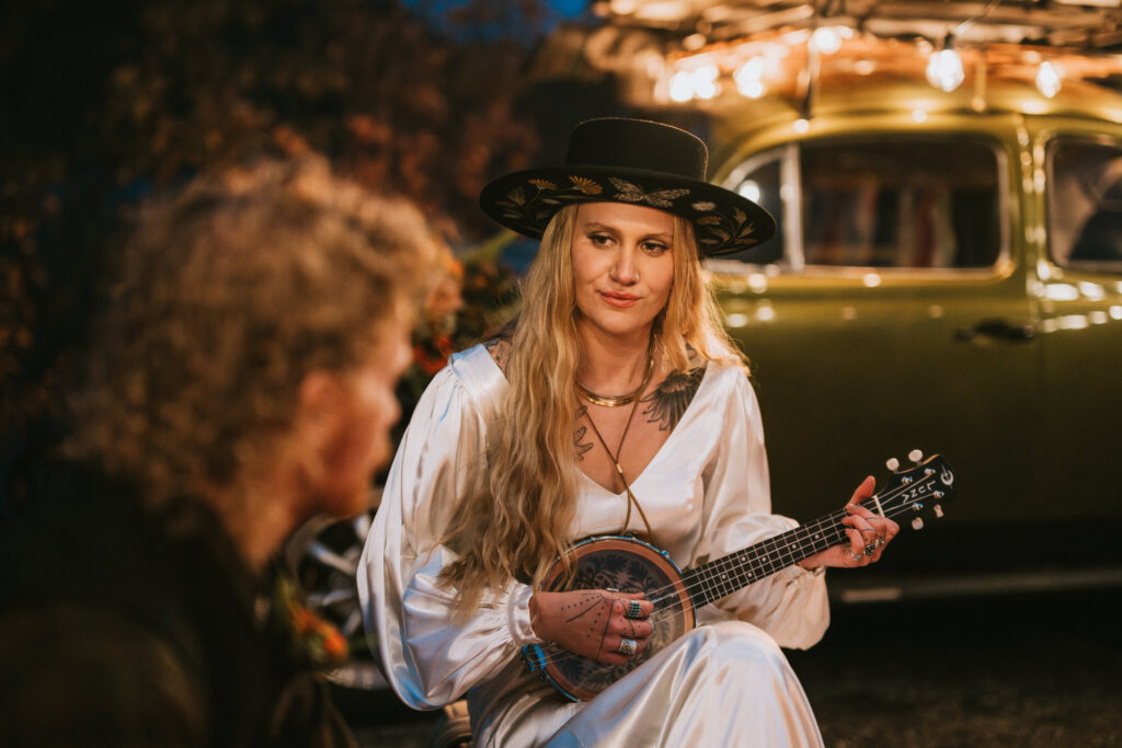 A woman in a white dress and black hat plays a string instrument while sitting outdoors. A person faces her, and a vintage car is in the background.