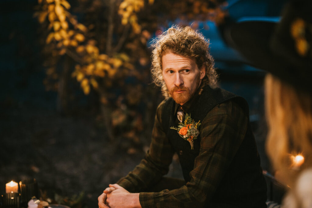 A person with curly hair and a beard sits outdoors by candlelight, wearing a dark vest and a flower boutonniere. Autumn foliage is visible in the background.