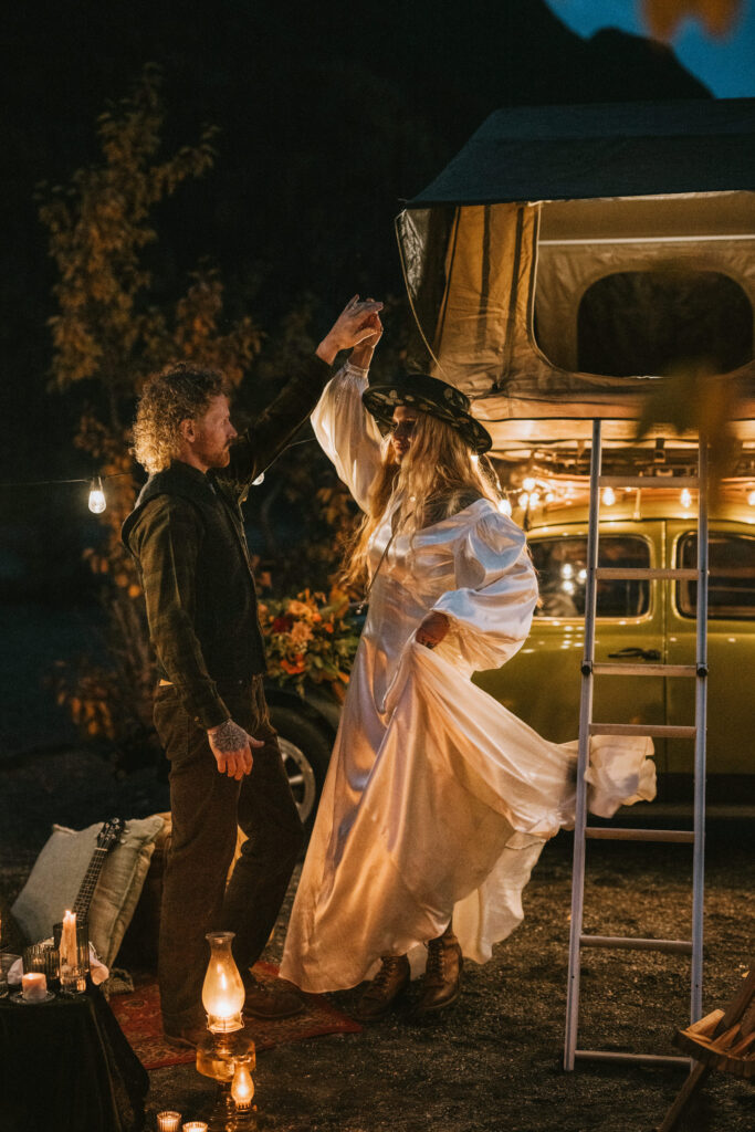 A man and woman dance outdoors at night near a lantern-lit campsite with a tent on top of a car.