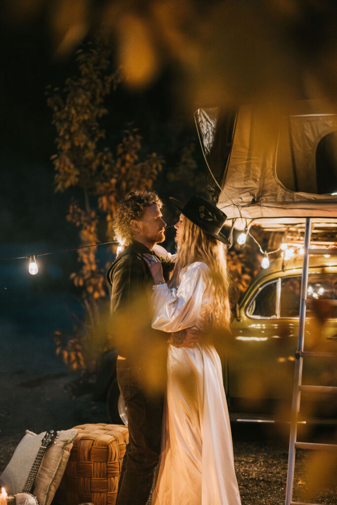 A couple stands close under string lights, near a vintage car with a rooftop tent. The scene is framed by autumn leaves, creating a cozy atmosphere.