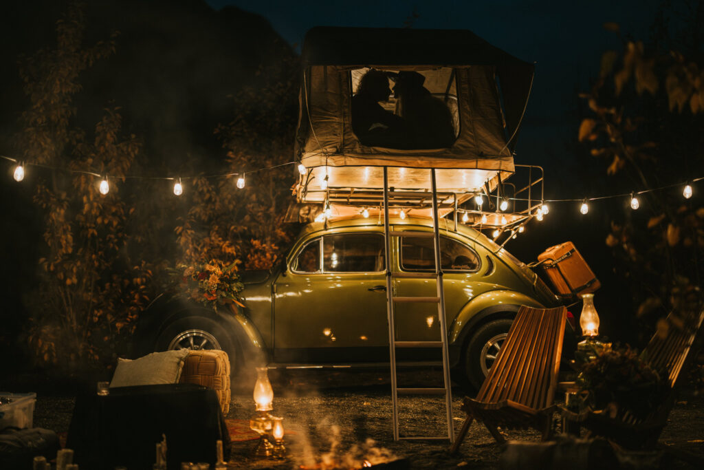 A vintage car with a rooftop tent is parked at night, surrounded by string lights and camping chairs. A warm fire burns nearby under a starry sky.