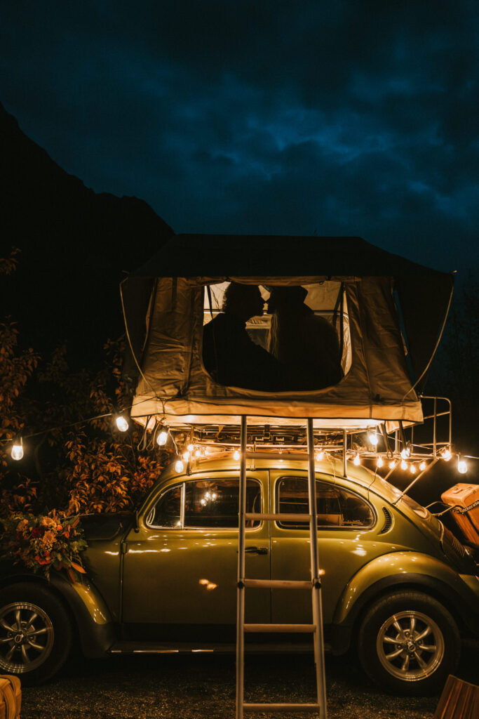 A vintage car with a rooftop tent and string lights. Two people sit inside the tent at dusk, with a backdrop of trees and a dark sky.