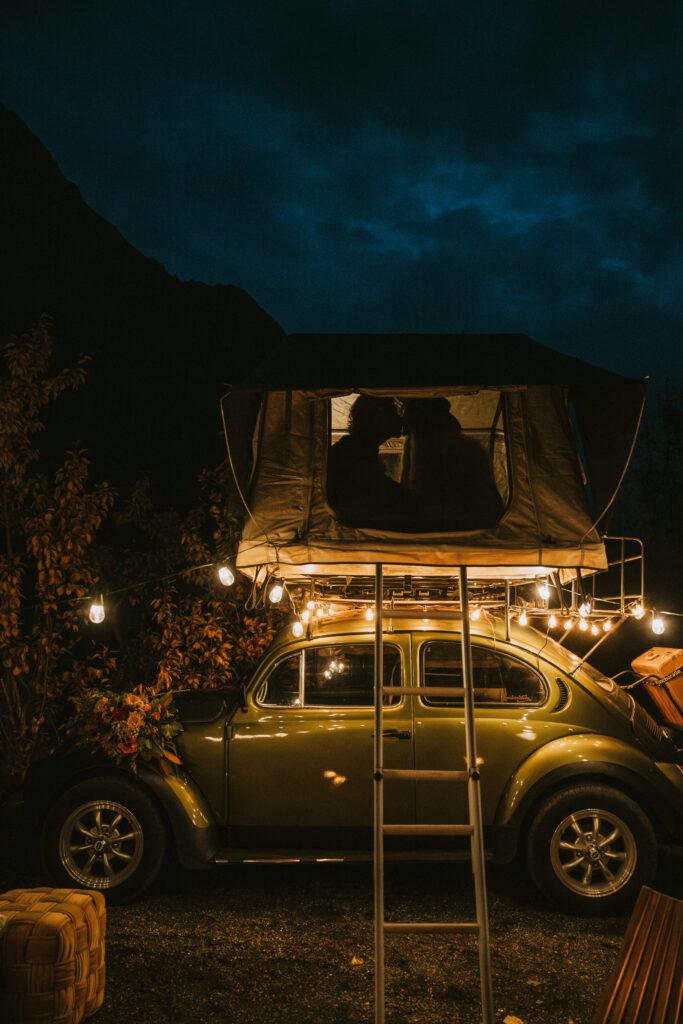 A green vintage car with a rooftop tent is illuminated by string lights at night. Two silhouetted figures are inside the tent.