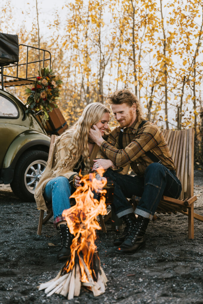 A couple sits by a campfire, surrounded by autumn trees, with an old car in the background.