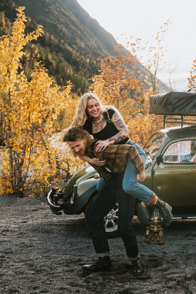 A person gives another a piggyback ride in front of a vintage car with autumn trees and mountains in the background.