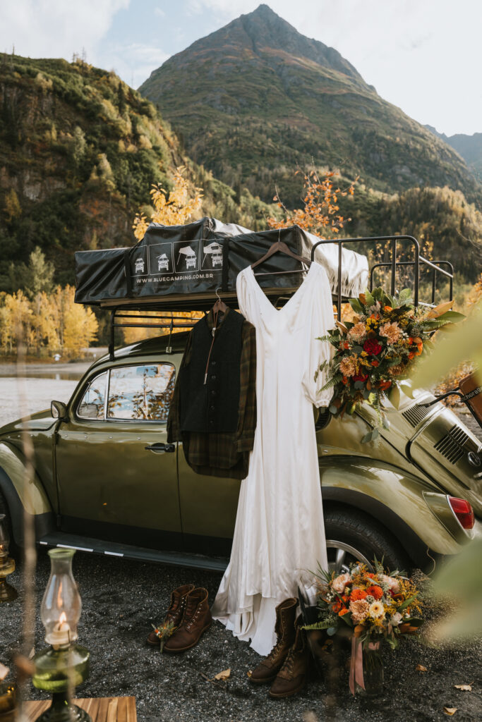 A vintage car with a roof tent is parked by a lake and mountain, displaying a white dress and coat. Brown boots and a bouquet of flowers are on the ground nearby for a fall camping elopement.