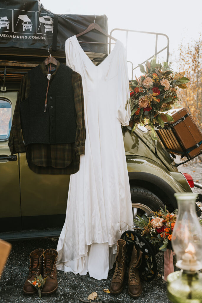 Wedding dress and bridal bouquet hanging outside a vintage car with boots and a plaid shirt, creating a rustic setting.