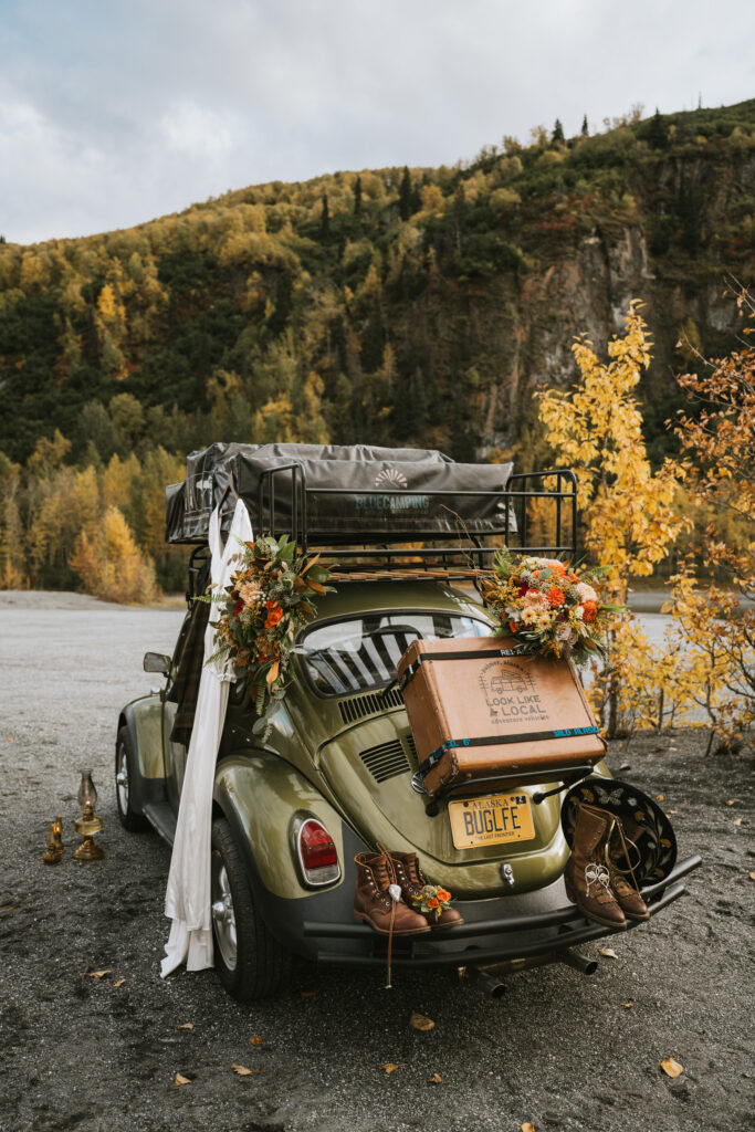 A vintage green car equipped with a roof tent and decorated with flowers is parked on a gravel path. Autumn trees and mountain cliffs are in the background.