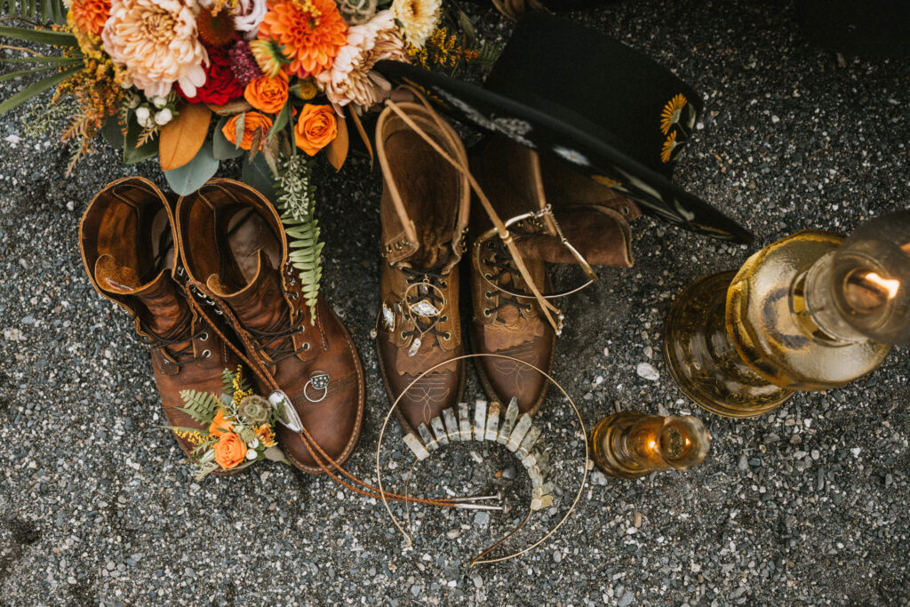 Brown boots, a necklace, and a hat are arranged on a gravel surface. Nearby, there are two lanterns and a bouquet of orange flowers.