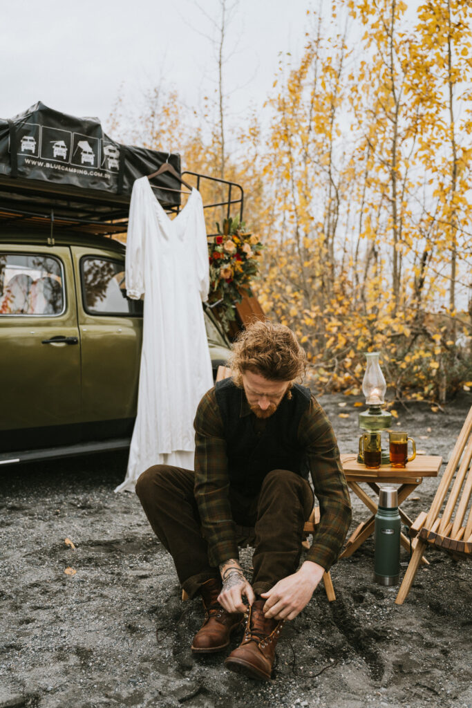 A person tying boots sits beside a green vehicle with a rooftop tent and hanging wedding dress. There are fall trees in the background and camping gear nearby.