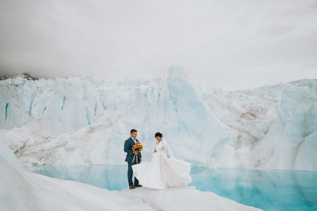 A couple in wedding attire stands on icy terrain beside a turquoise glacial lake, surrounded by vast glacier formations under a cloudy sky, epitomizing the magic of an adventurous destination wedding. Ensuring this dream moment is preserved, they smartly opted for destination wedding insurance.