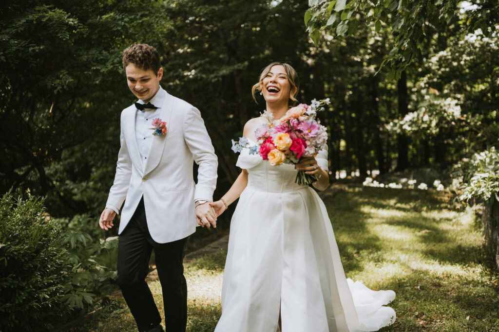 A bride and groom, covered by destination wedding insurance, walk hand in hand outdoors, the bride holding a colorful bouquet.