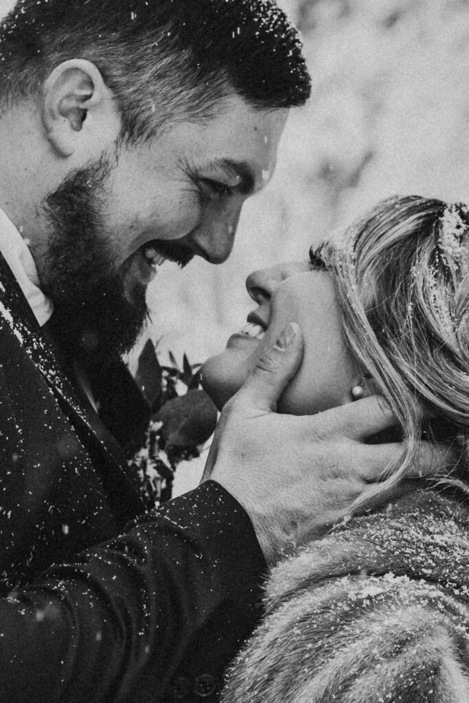 A couple smiling closely at each other in the snow, with their faces close together and the man's hand gently holding the woman's face.