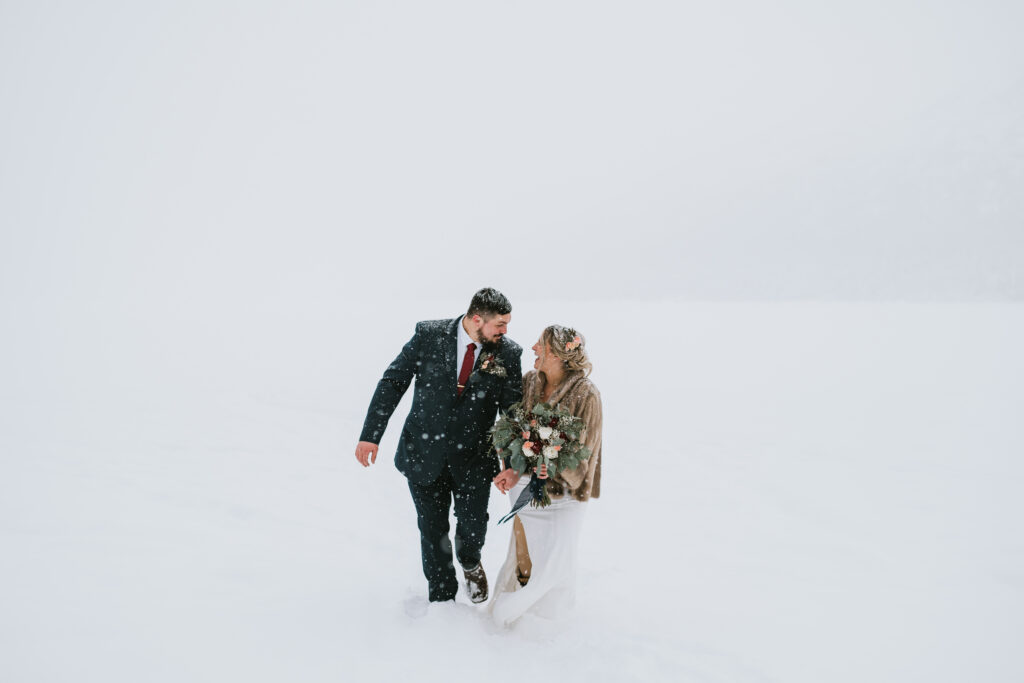 A couple dressed in formal attire strolls through a snowy landscape, the man in a dark suit and the woman in a white dress with a shawl, holding a bouquet. 