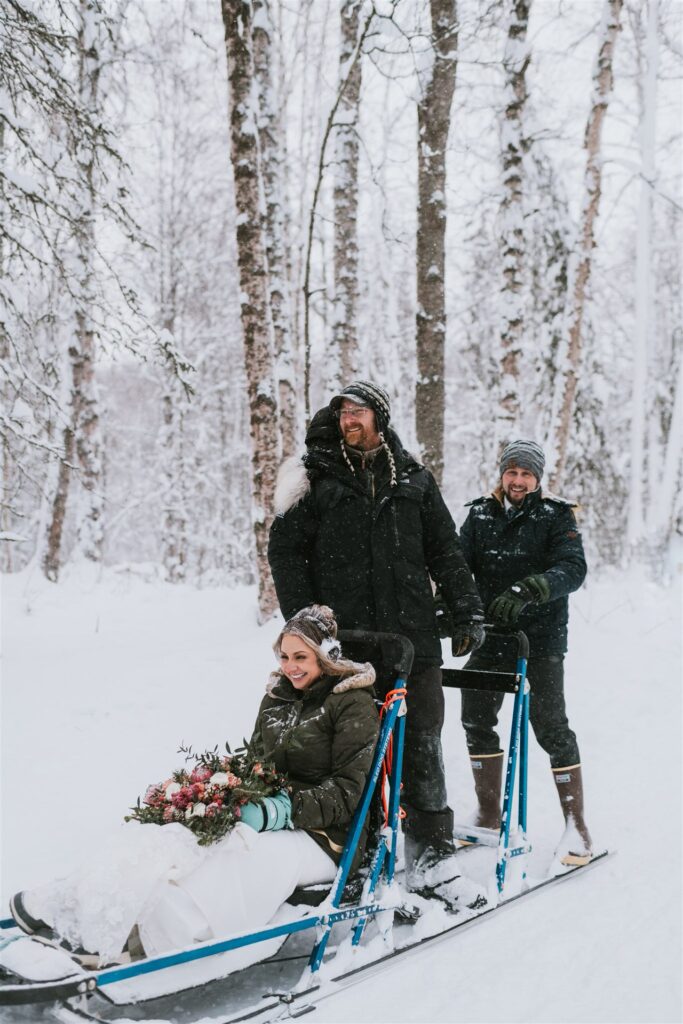 In a snowy winter forest, a person sits on a sled holding a bouquet, embodying the spirit of an impromptu winter elopement, while two others stand behind them, all wrapped warmly in their winter clothing.