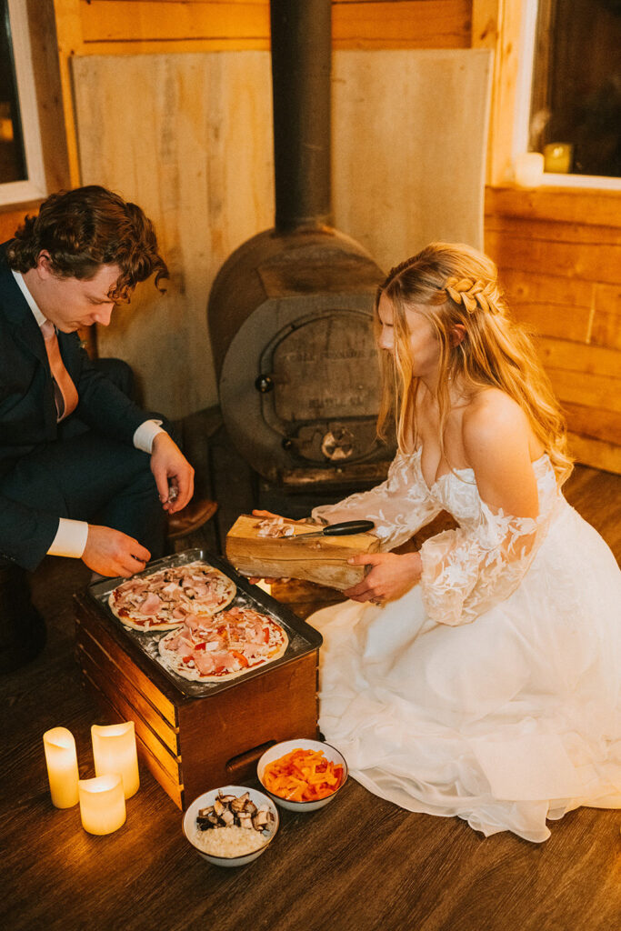 In a cozy setting perfect for a winter elopement, the bride and groom prepare to cook pizzas on a wood-fired stove. The room glows warmly with candles on the floor, while bowls of toppings sit invitingly nearby.