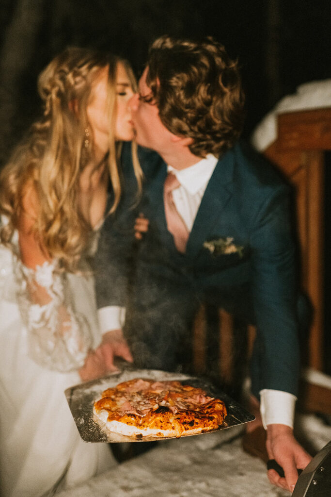 A couple in formal attire shares a kiss while holding a pizza on a tray, capturing the warmth and spontaneity of their winter elopement.