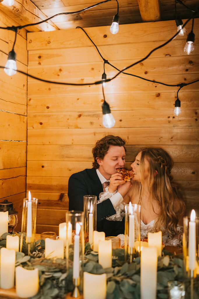 A couple in formal attire sits in a cozy, wooden booth sharing food, celebrating their winter elopement. Candles and string lights create a warm ambiance.