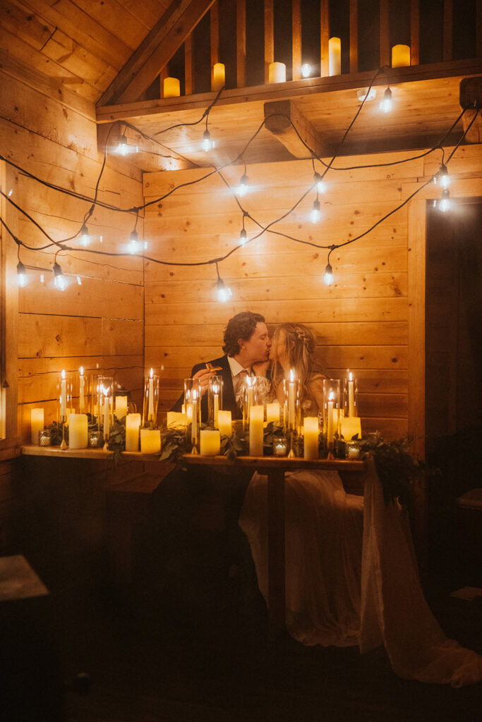 A couple sits at a candlelit wooden table in a cozy room, surrounded by string lights and numerous candles, creating a warm, romantic atmosphere perfect for an intimate winter elopement.