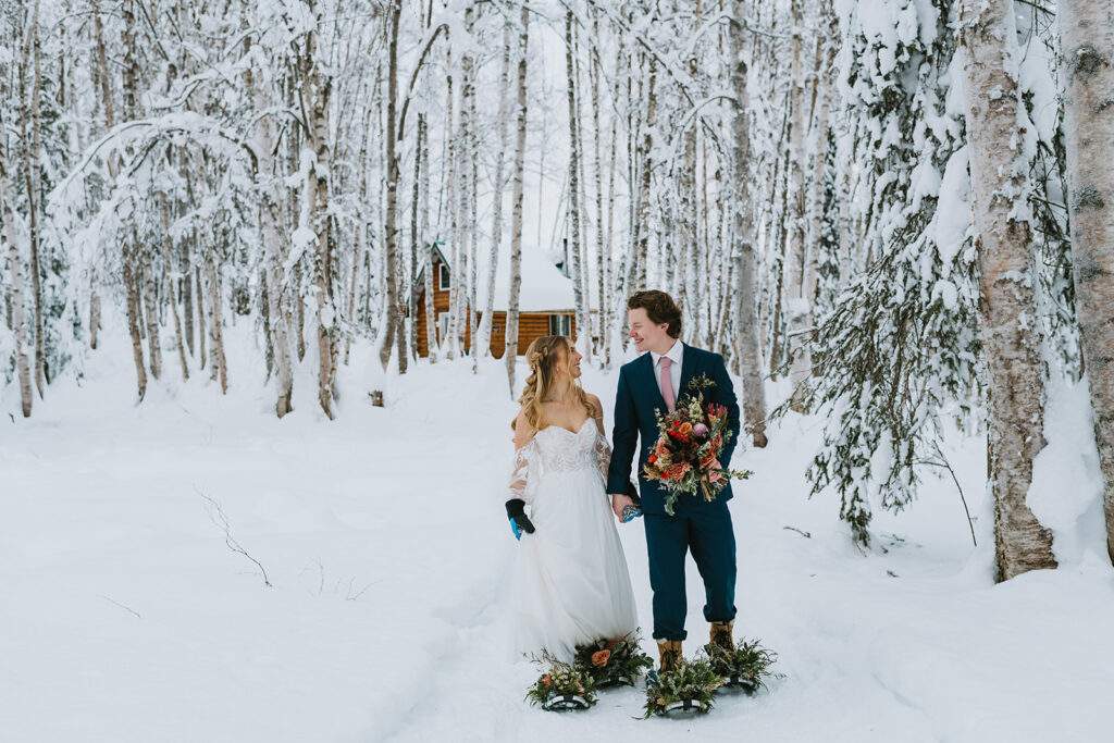 A bride and groom stand on a snow-covered path in a forest, holding a bouquet, embracing the magic of a winter elopement with a rustic cabin in the background.