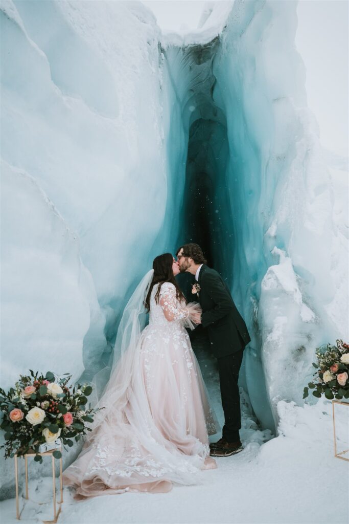 A couple kisses in front of an ice cave during their winter elopement. The bride wears a flowing gown, and the groom is in a suit. Snow blankets the ground, with flower arrangements on either side.