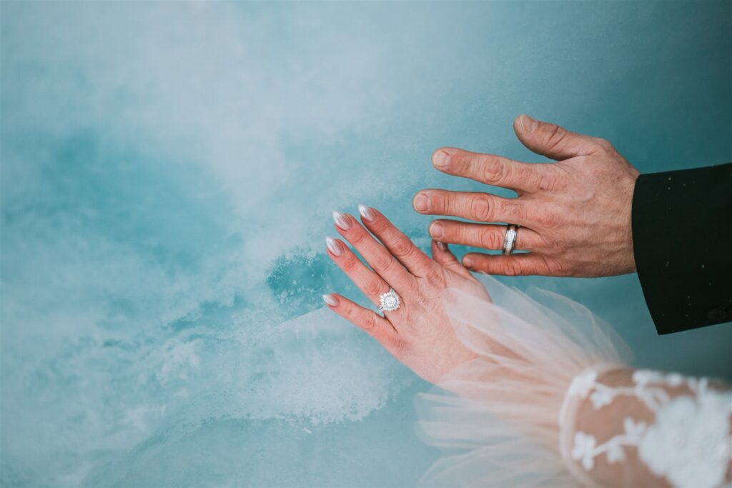 Two hands with wedding rings rest against a serene blue background. The left hand features intricate bridal lace detailing, evoking the delicate beauty of a winter elopement.