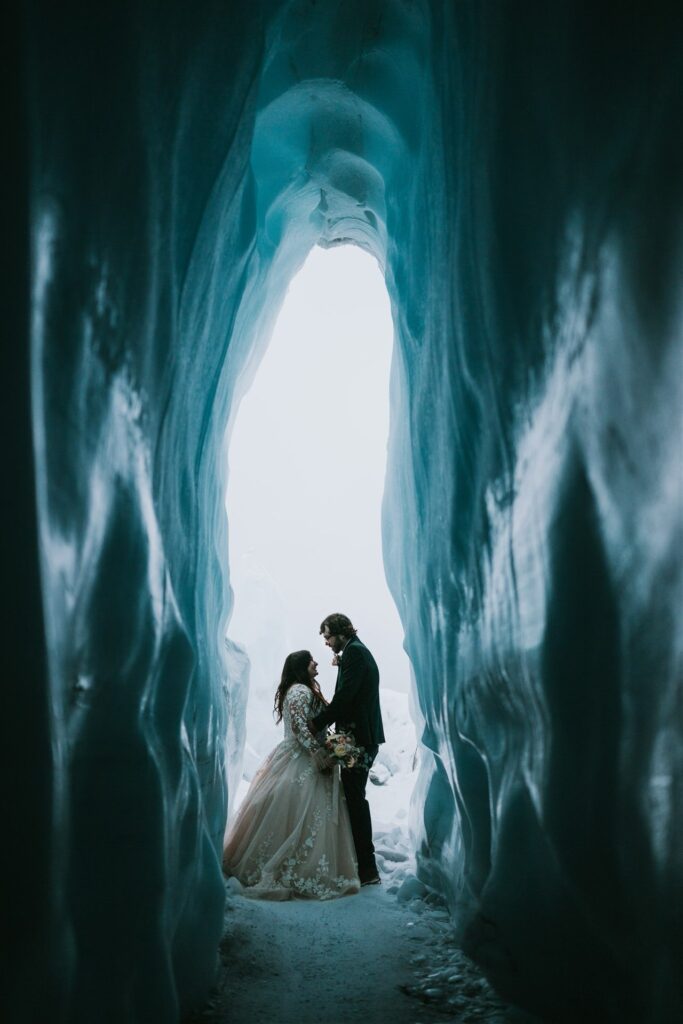 A couple stands together in a narrow ice cave passage, the soft light from their winter elopement illuminating them from behind.