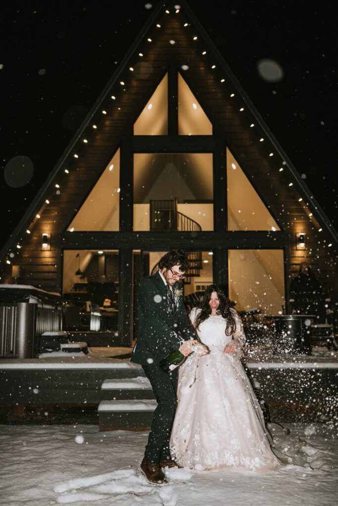 Bride and groom joyfully celebrating their winter elopement in front of a lit A-frame cabin at night, with snow gently falling around them.