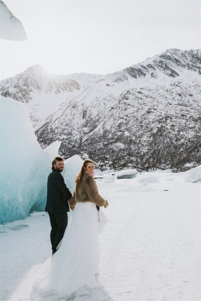 A couple in wedding attire stands on a snowy landscape with large ice formations and mountains in the background, facing forward and holding hands during their intimate winter elopement.