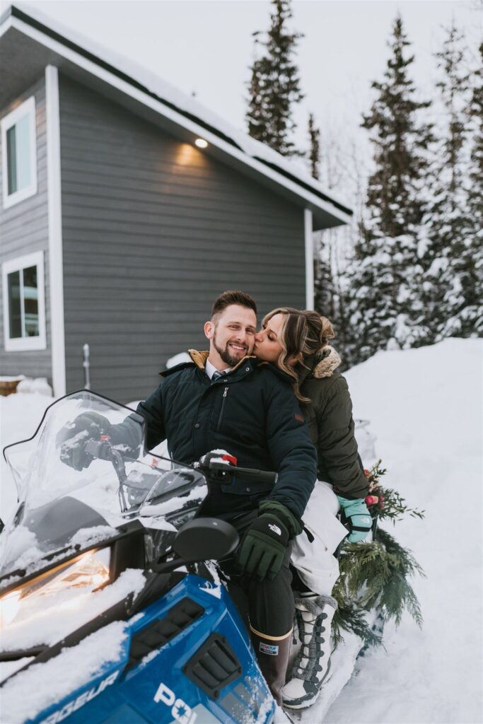 A couple enjoys a cozy winter elopement on a snowmobile, the woman playfully planting a kiss on the man's cheek. In the background, a gray house and towering evergreen trees complete the serene snowy landscape.