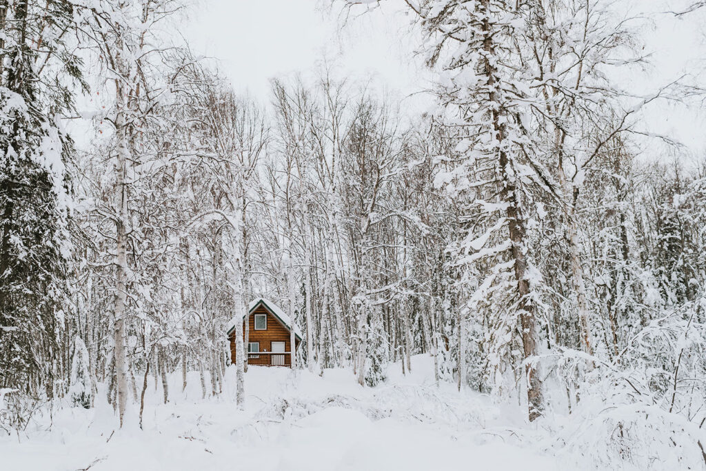 A small wooden cabin stands surrounded by snow-covered trees in a winter forest landscape, making it the perfect setting for an intimate winter elopement.