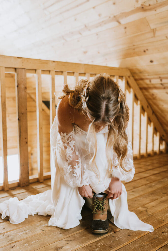 Amidst a cozy, rustic setting, a bride in a white lace wedding dress bends down to tie her lace-up boots on the wooden floor, preparing for an intimate winter elopement.