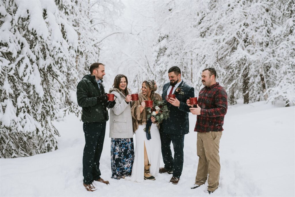 A group of people in winter clothing gather in a snowy forest, red mugs in hand, celebrating a cozy winter elopement. One person holds a bouquet of flowers, adding a touch of romance to the chilly scene.