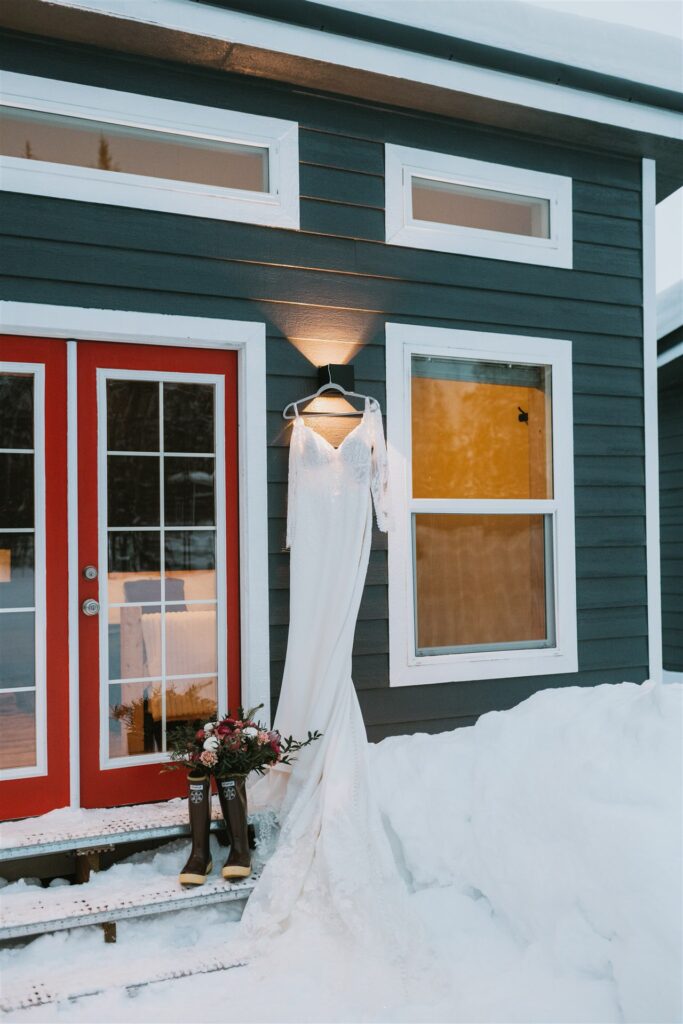 A long white wedding dress hangs outside a house with red doors, next to a bouquet of flowers and a pair of boots. Snow covers the ground, setting the perfect scene for a winter elopement.