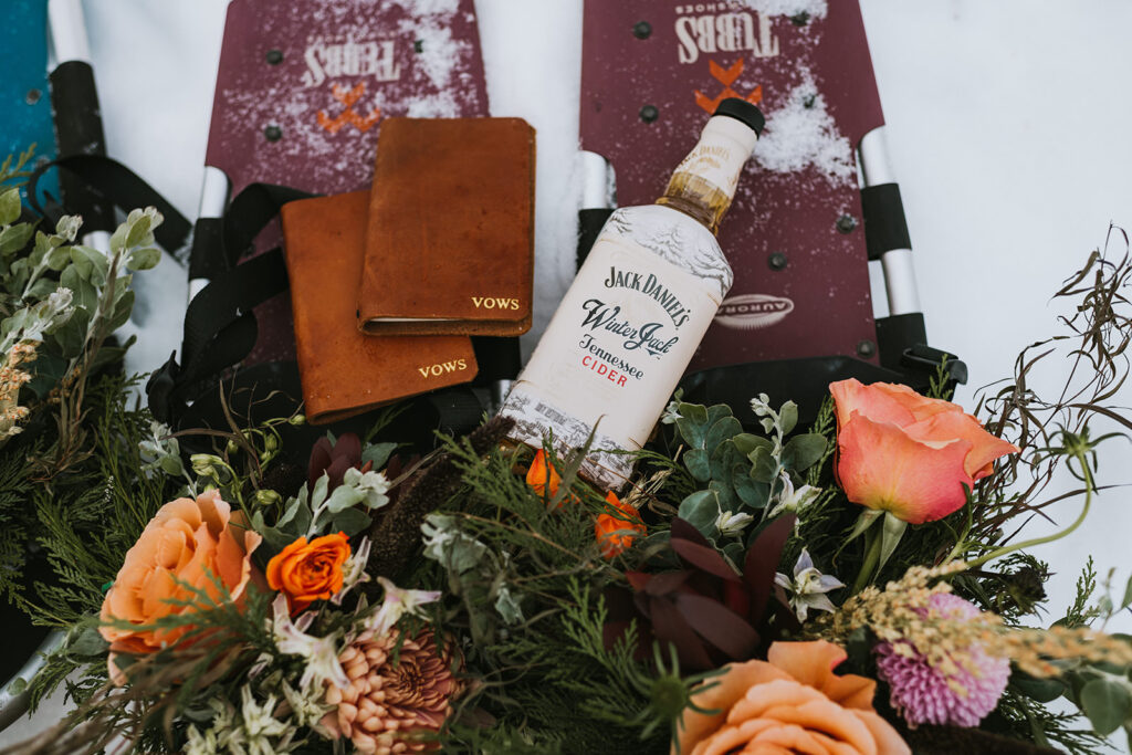 A bottle of Jack Daniel's Winter Jack sits among vow books and bouquets of flowers, while snow-covered snowshoes lay elegantly on the snow, capturing the essence of a magical winter elopement.